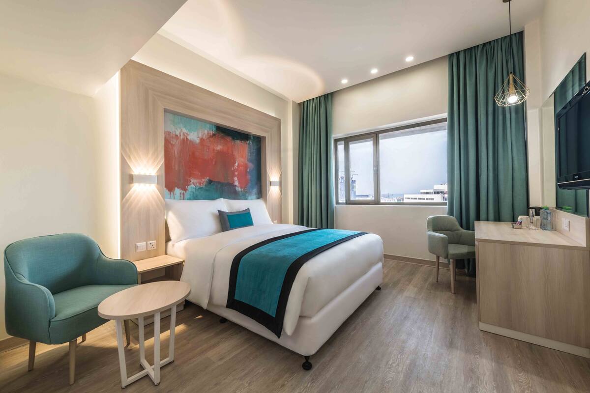 A modern hotel room with a bed, a desk, a chair, and a view of the city.