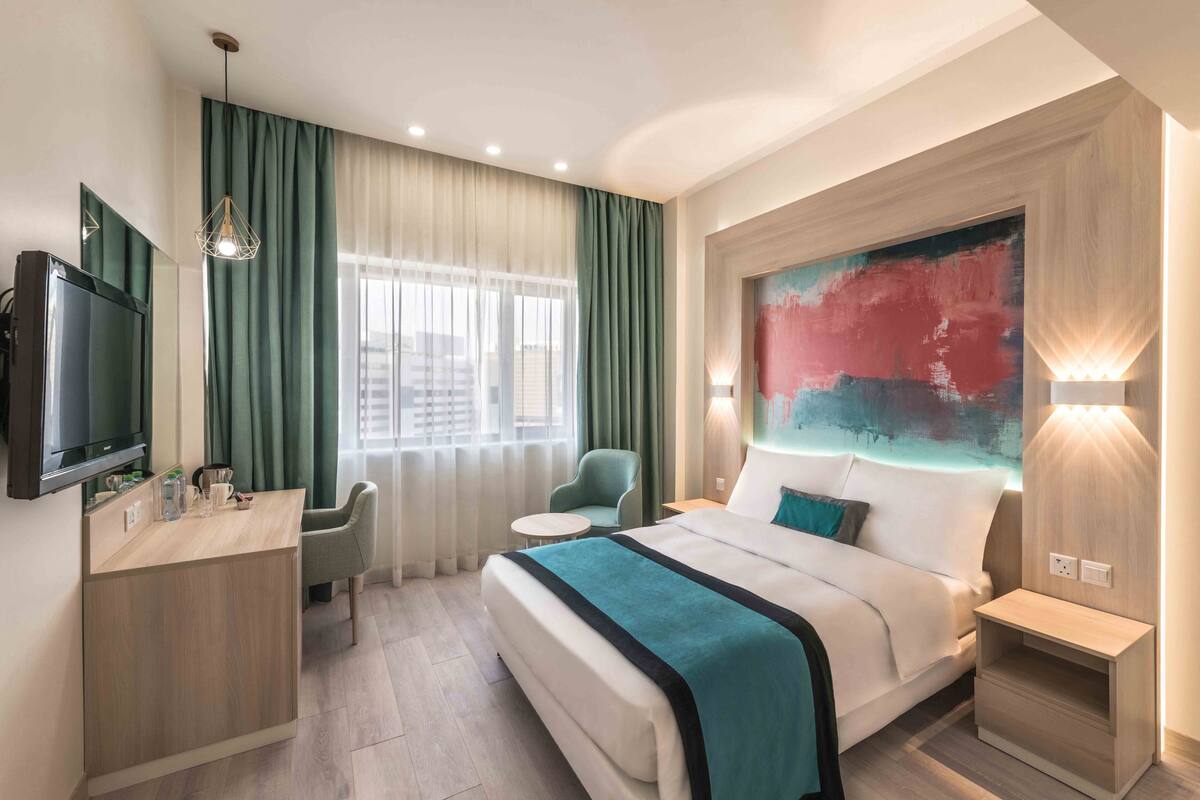 A hotel room with a bed, a desk, a chair, a TV, and a large abstract painting on the wall.