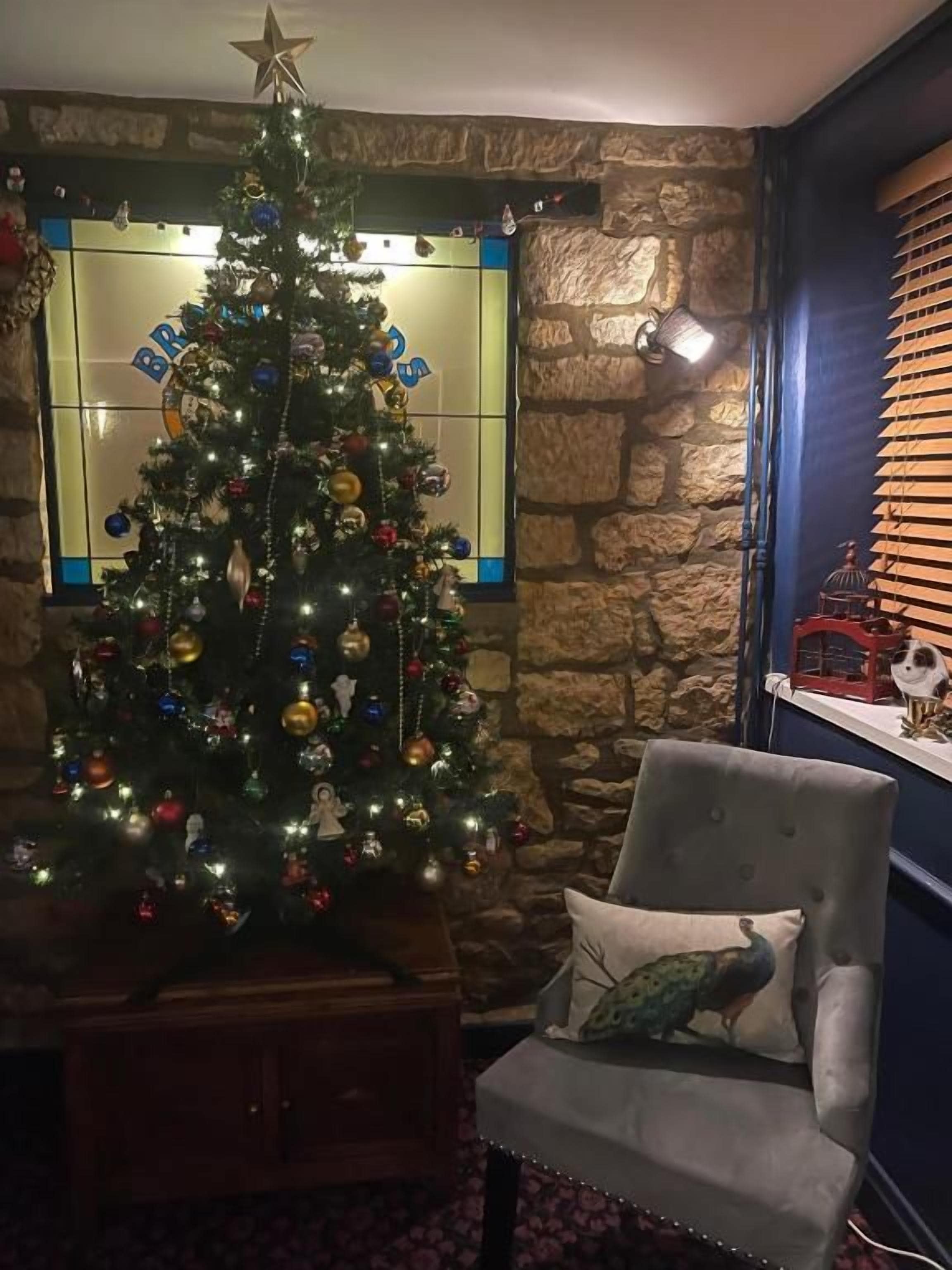 Un sapin de Noël placé dans un coin, avec un mur en pierre et une chaise agrémentée d’un coussin à motif de paon.