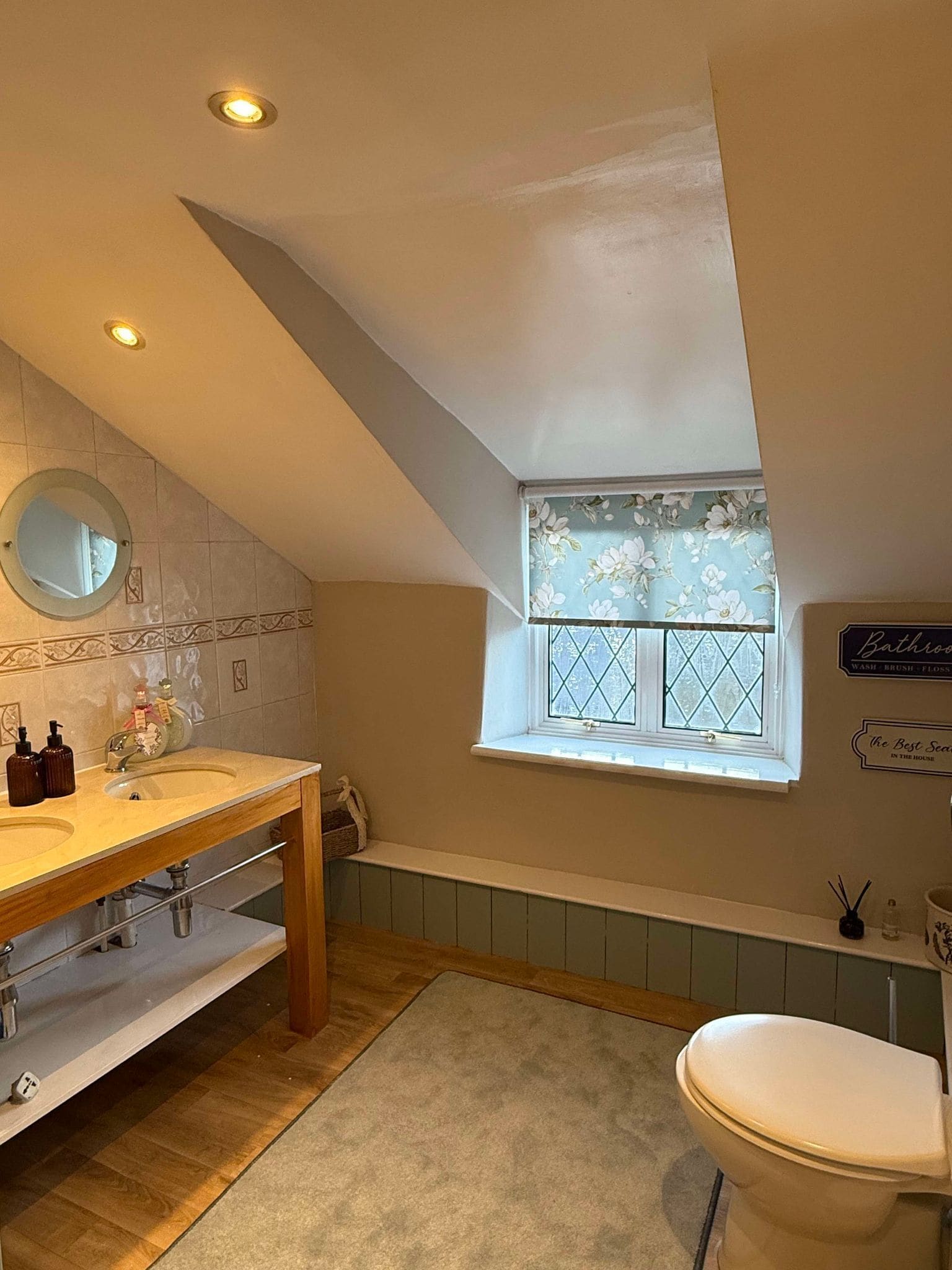 Une salle de bain avec un meuble-lavabo en bois, un miroir rond, une fenêtre ornée de rideaux à motifs floraux et des toilettes.