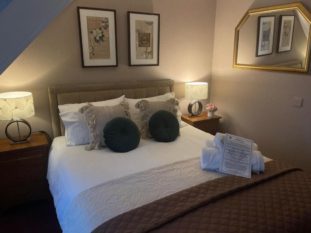 Habitación de hotel con cama, almohadas, mesita de noche con lámpara, un cuadro enmarco y un espejo.