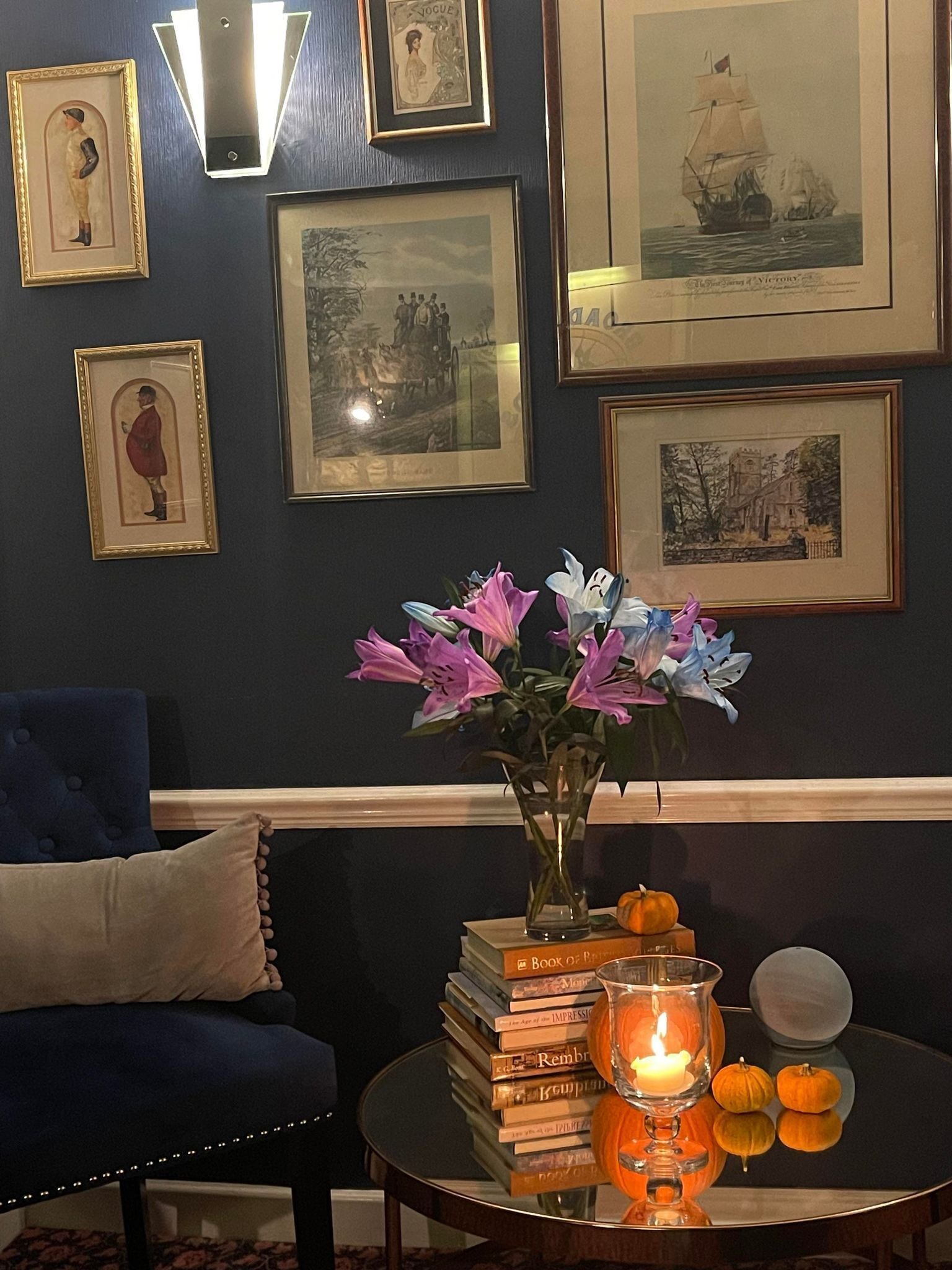 A room with a blue sofa, a round table with books, a vase with flowers, a candle, and framed pictures on the wall.