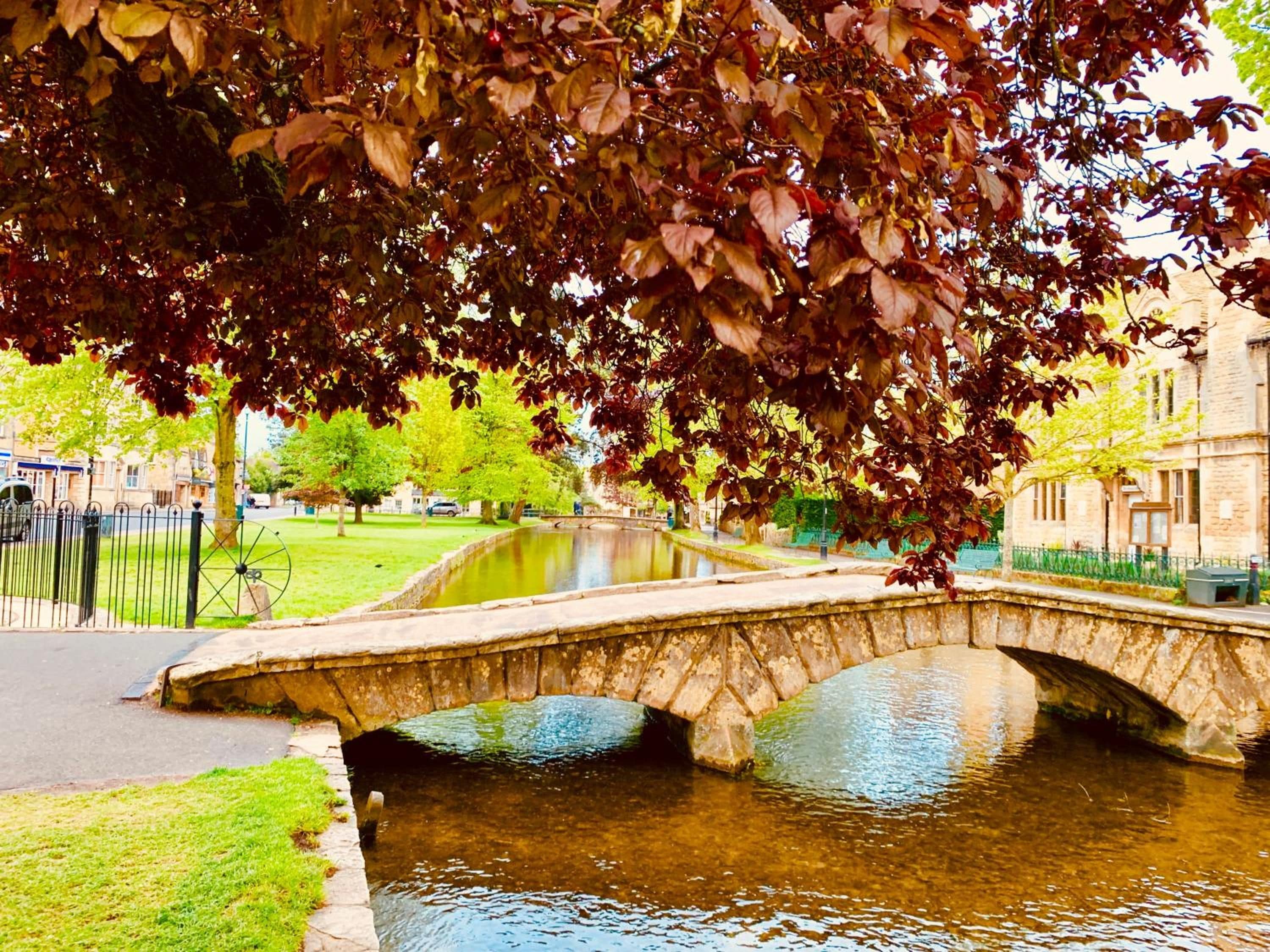 A stone bridge over a calm water body, surrounded by greenery and buildings.