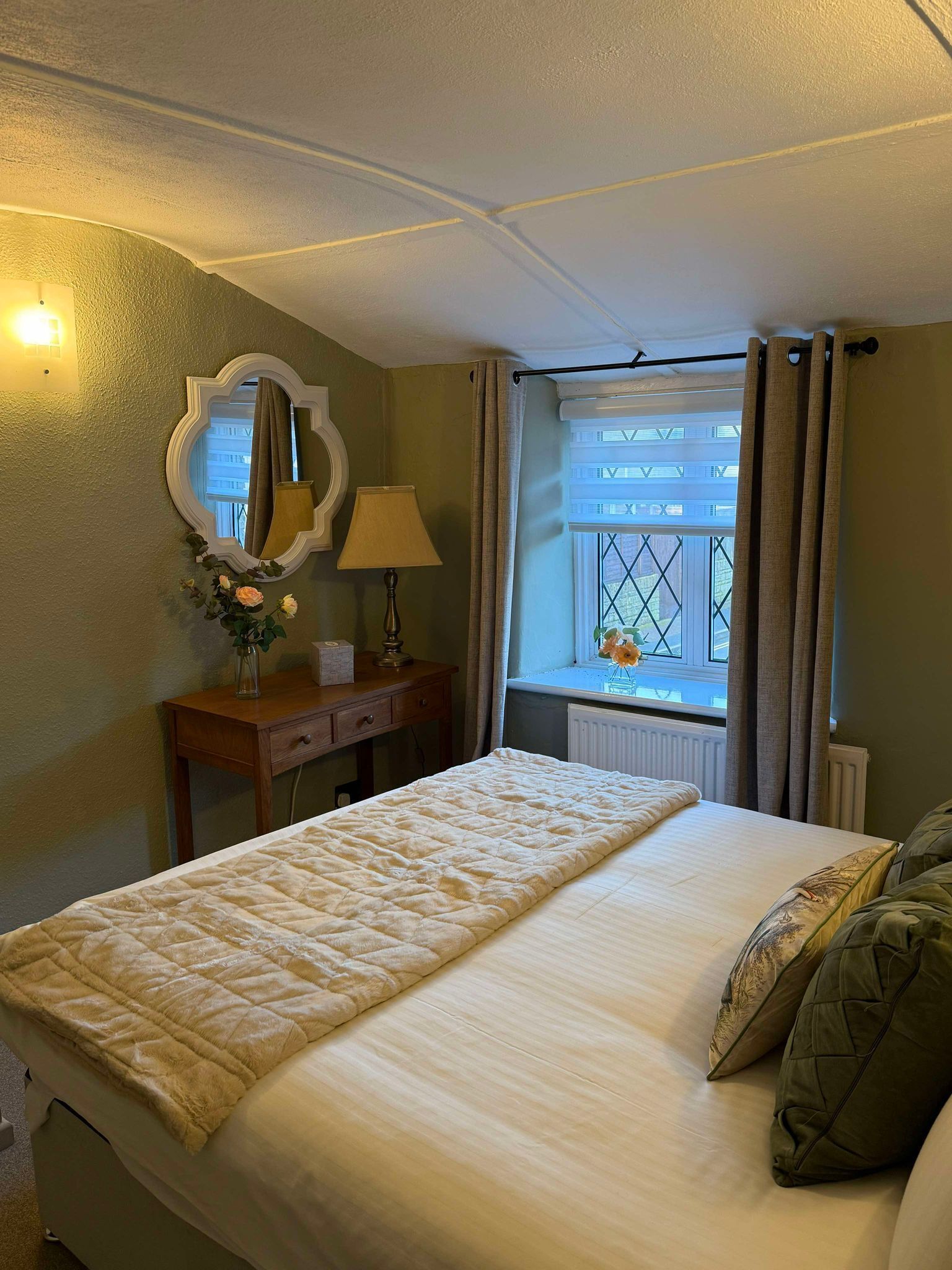 Un lit bien fait, recouvert d’une couverture matelassée, une table de chevet avec une lampe et un vase de fleurs, un miroir au mur et une fenêtre avec des rideaux.
