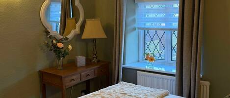 Un lit bien fait, recouvert d’une couverture matelassée, une table de chevet avec une lampe et un vase de fleurs, un miroir au mur et une fenêtre avec des rideaux.