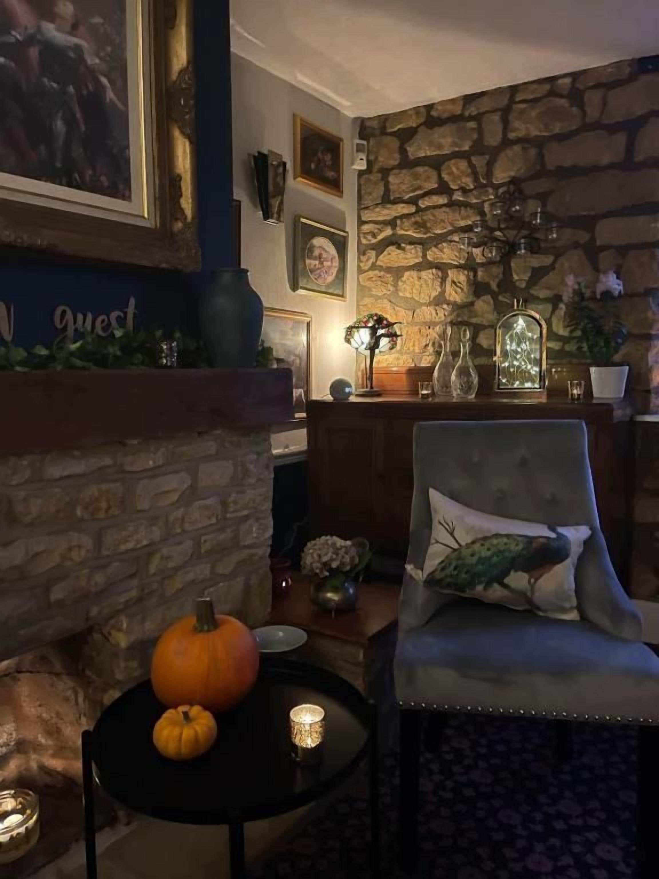 A cozy room with a stone fireplace, a round table with pumpkins, a chair with a decorative pillow, and a wall with framed pictures.