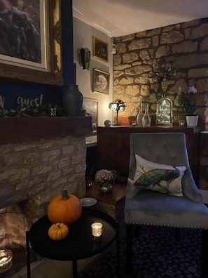 Une pièce chaleureuse avec une cheminée en pierre, une table ronde décorée de citrouilles, un fauteuil agrémenté d’un coussin décoratif et un mur orné de tableaux encadrés.