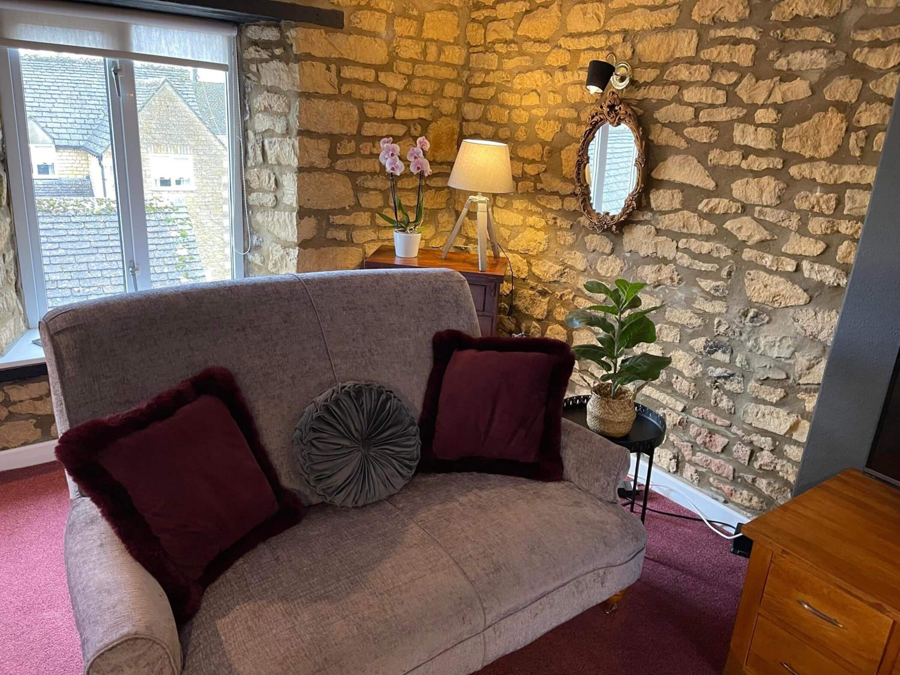 A cozy room with a stone wall, a cushioned sofa with two pillows, a lamp, a mirror, and a potted plant.