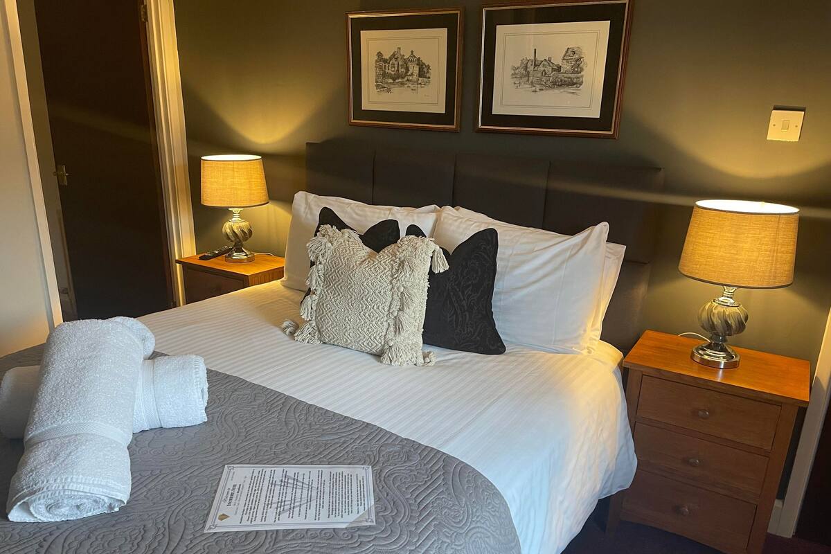 A neatly made bed with a patterned bedspread, two bedside lamps, a nightstand with a lamp, and two framed pictures on the wall.
