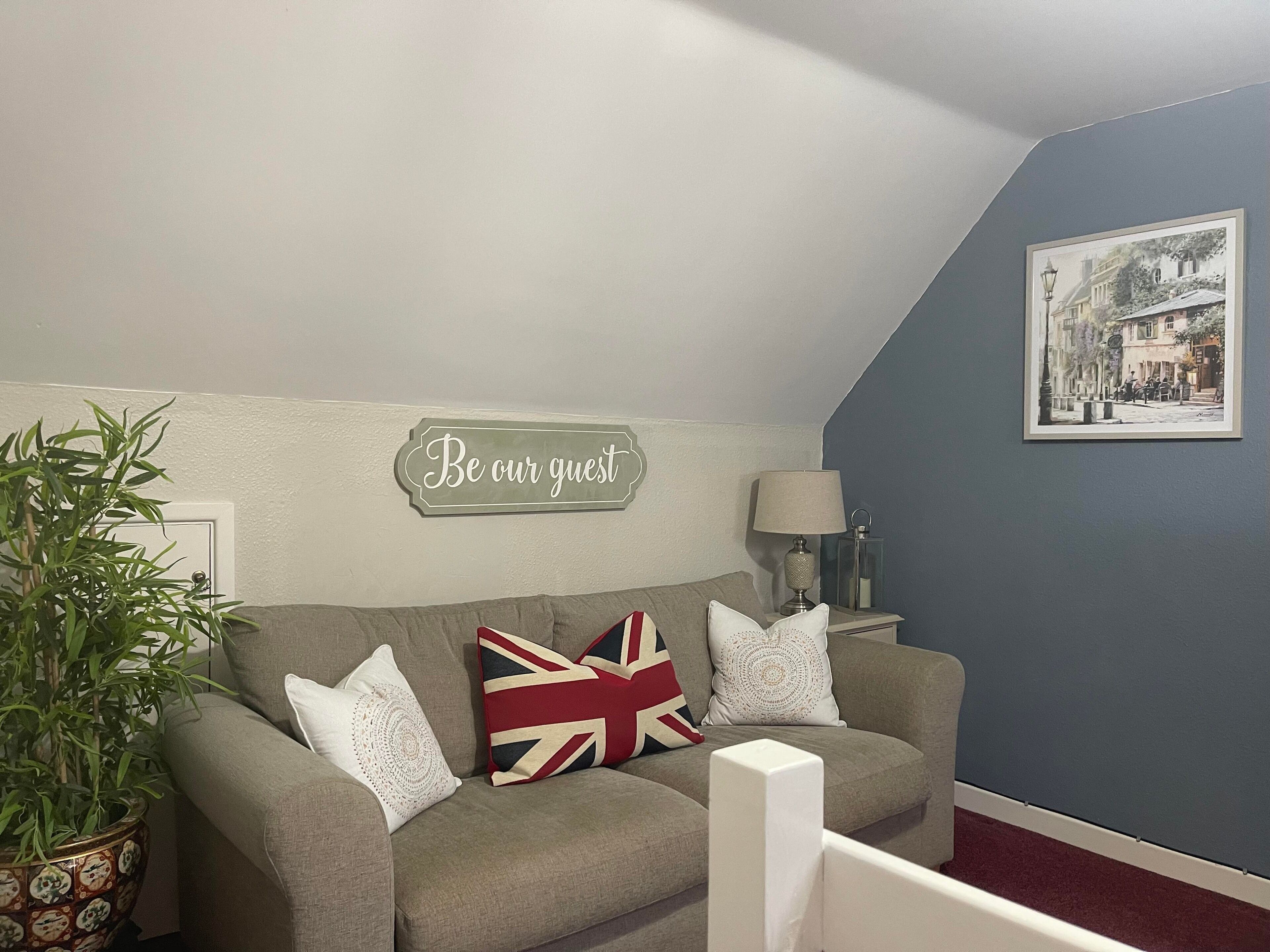 A cozy living room with a grey sofa, a 'Be our guest' sign, a lamp, and a framed picture on the wall.