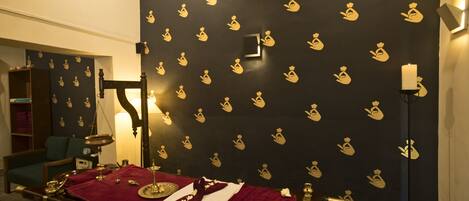 A room with a massage table, a dark wall with golden patterns, a lamp, and a bookshelf.