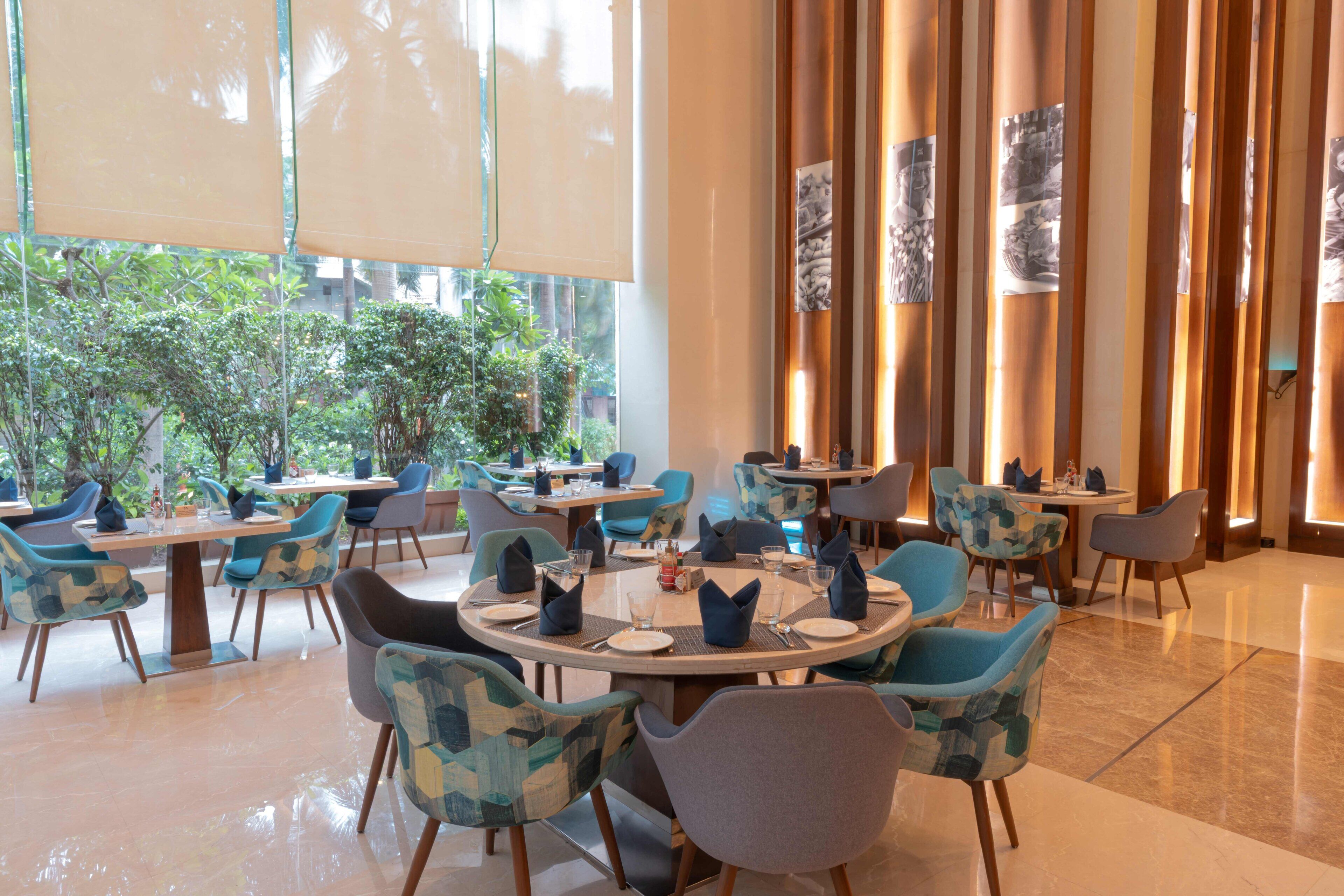 A modern dining area with round tables and chairs, large windows with blinds, and indoor plants.
