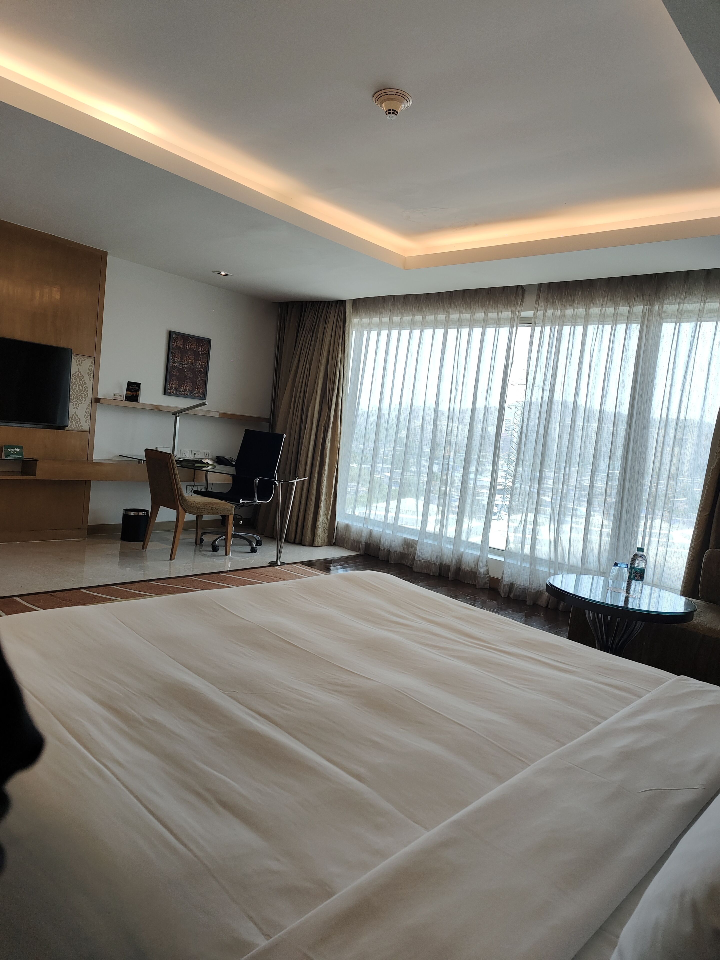 A hotel room with a large bed, a desk with a chair, a TV, and a view of the city through sheer curtains.