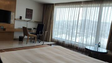 A hotel room with a large bed, a desk with a chair, a TV, and a view of the city through sheer curtains.