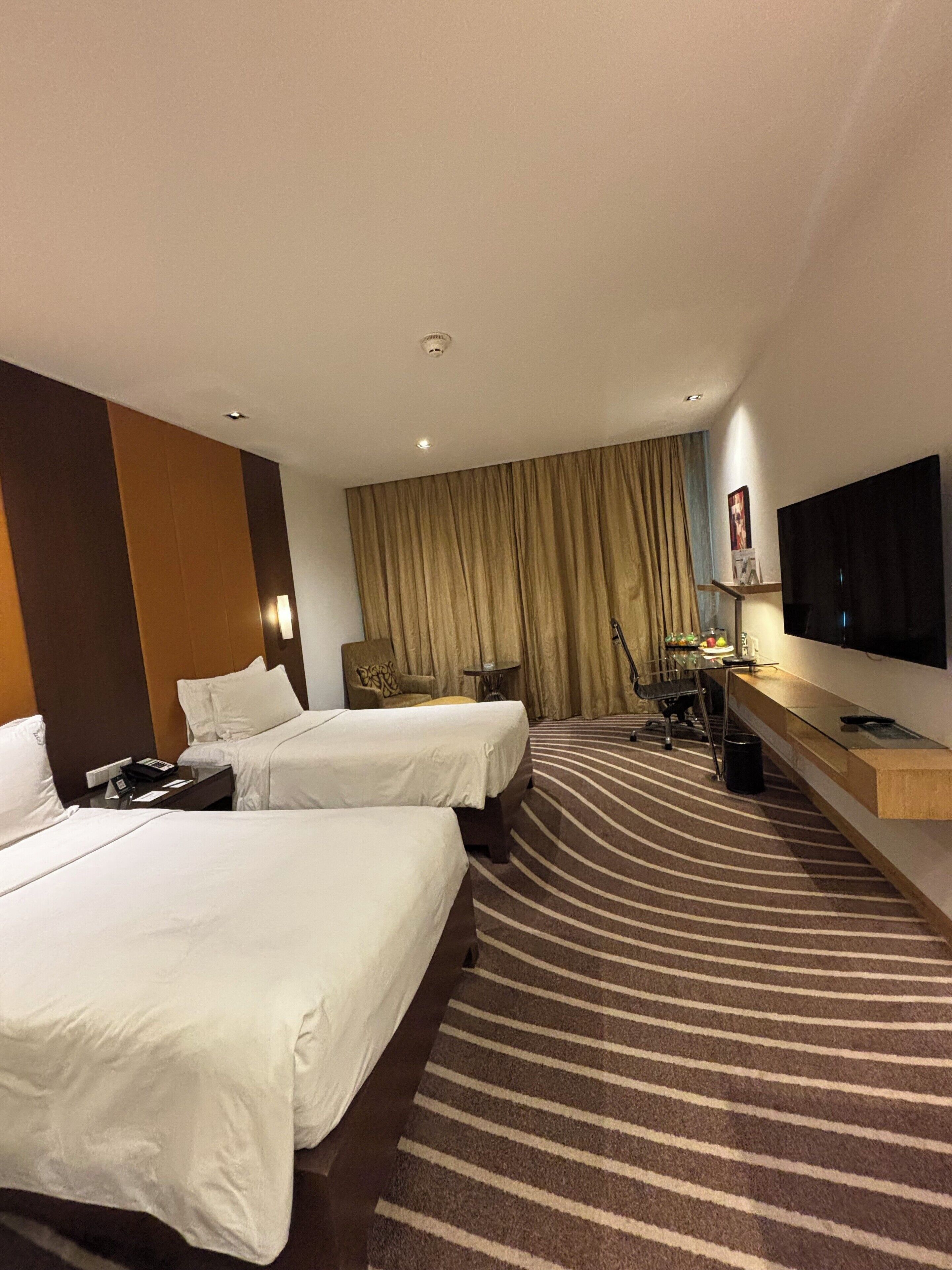 A hotel room with two beds, a desk with a flat-screen TV, a chair, a small table, and a large window with curtains.