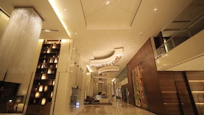 A modern hotel lobby with a high ceiling, a large chandelier, a reception desk, and a staircase.
