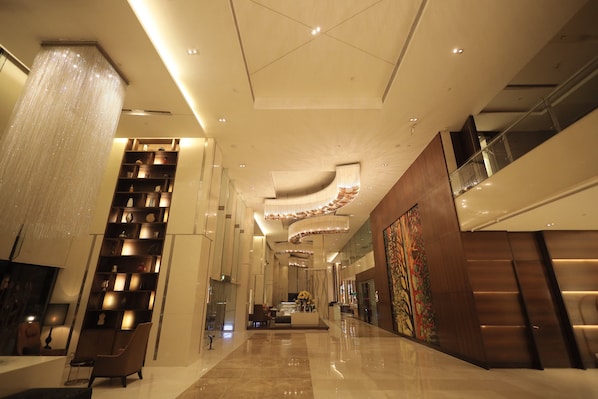 A modern hotel lobby with a high ceiling, a large chandelier, a reception desk, and a staircase.