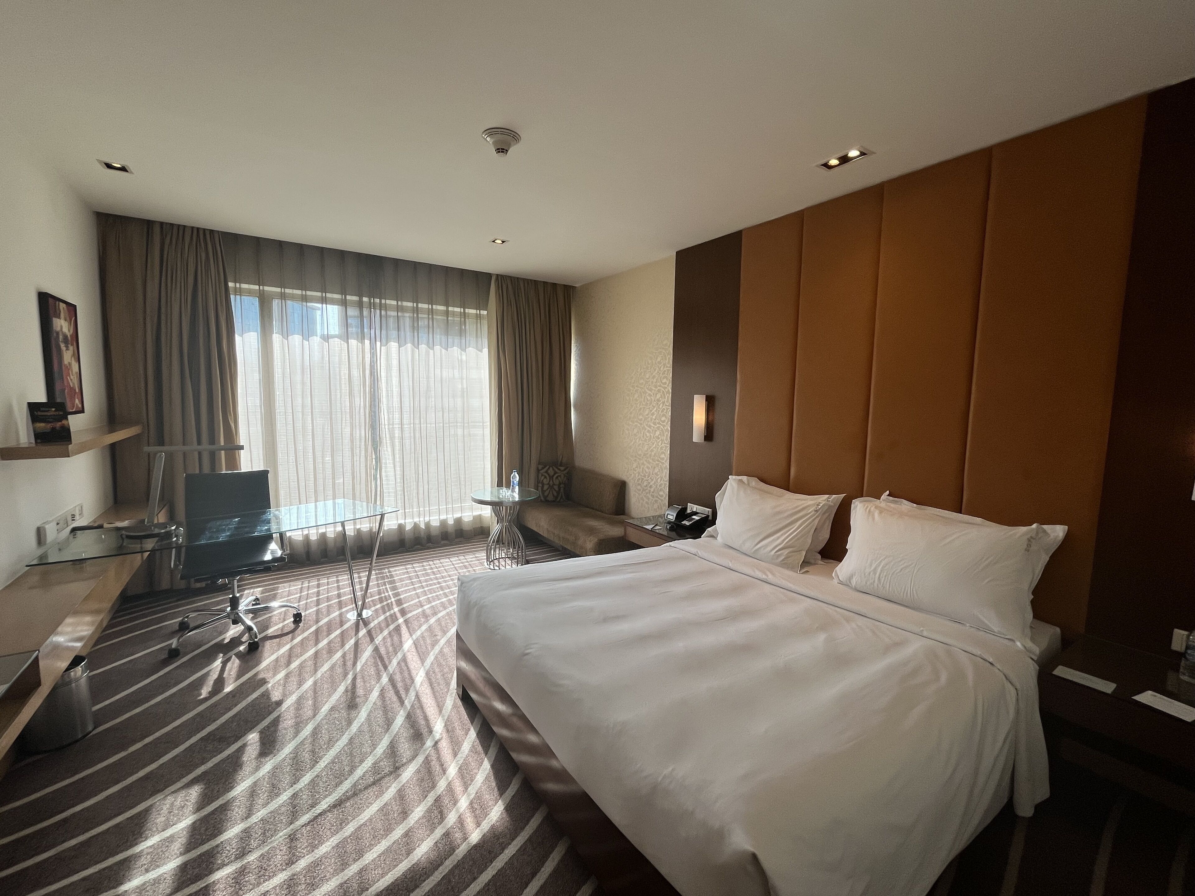 A hotel room with a large bed, a desk with a chair, a sofa, and a large window with curtains.