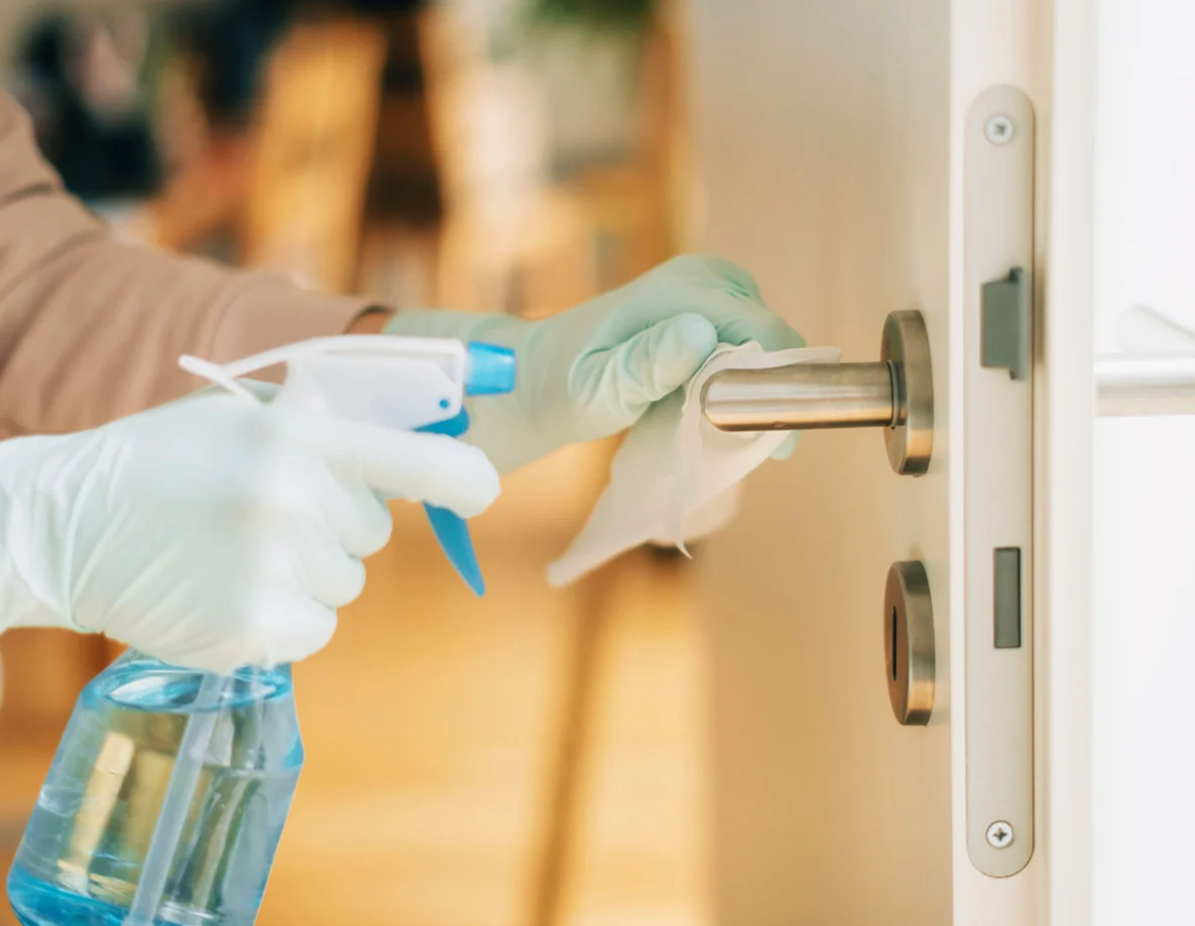 A person wearing gloves using a spray bottle to disinfect a door handle.