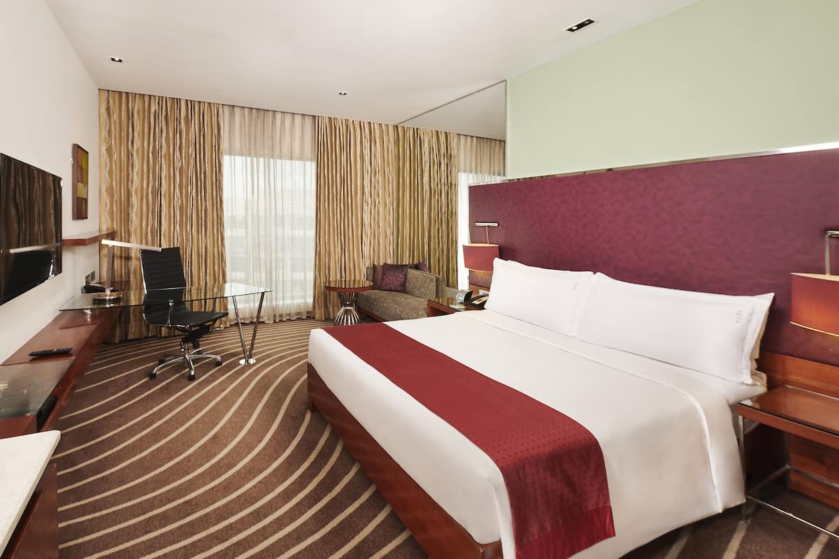 A hotel room with a large bed, a desk with a chair, a TV, and a large window with curtains.