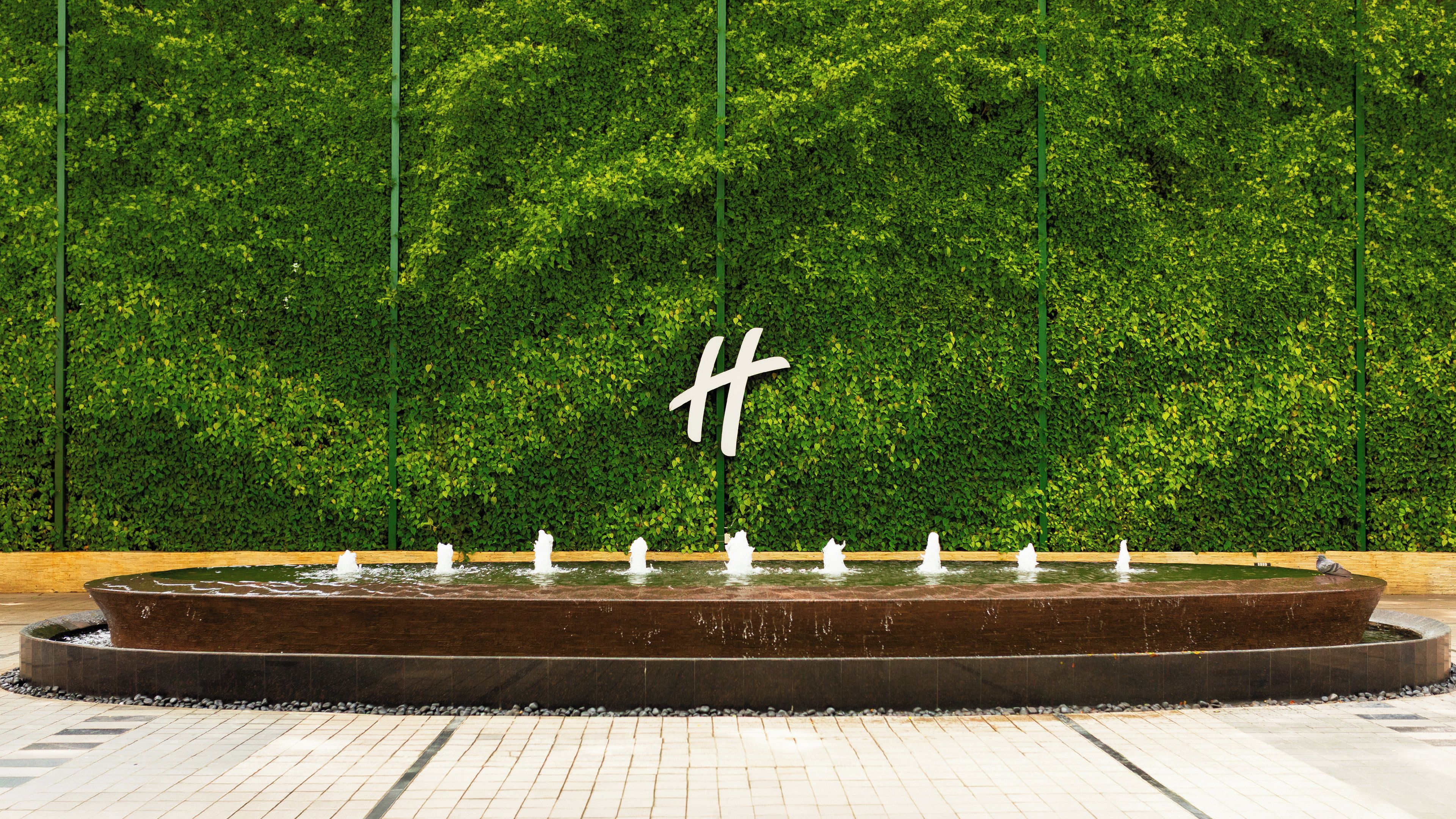 A circular water feature with fountains in front of a hedge wall with a large 'H' sign.