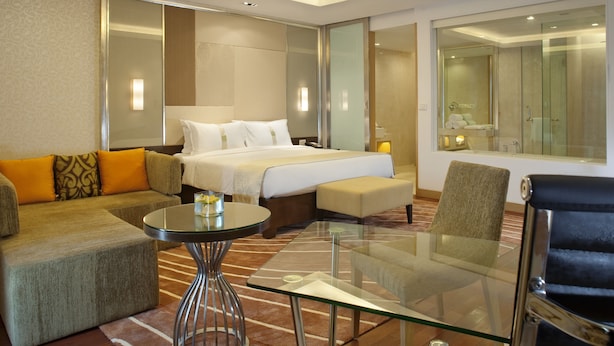 A modern hotel room with a bed, sofa, armchair, and a glass table.