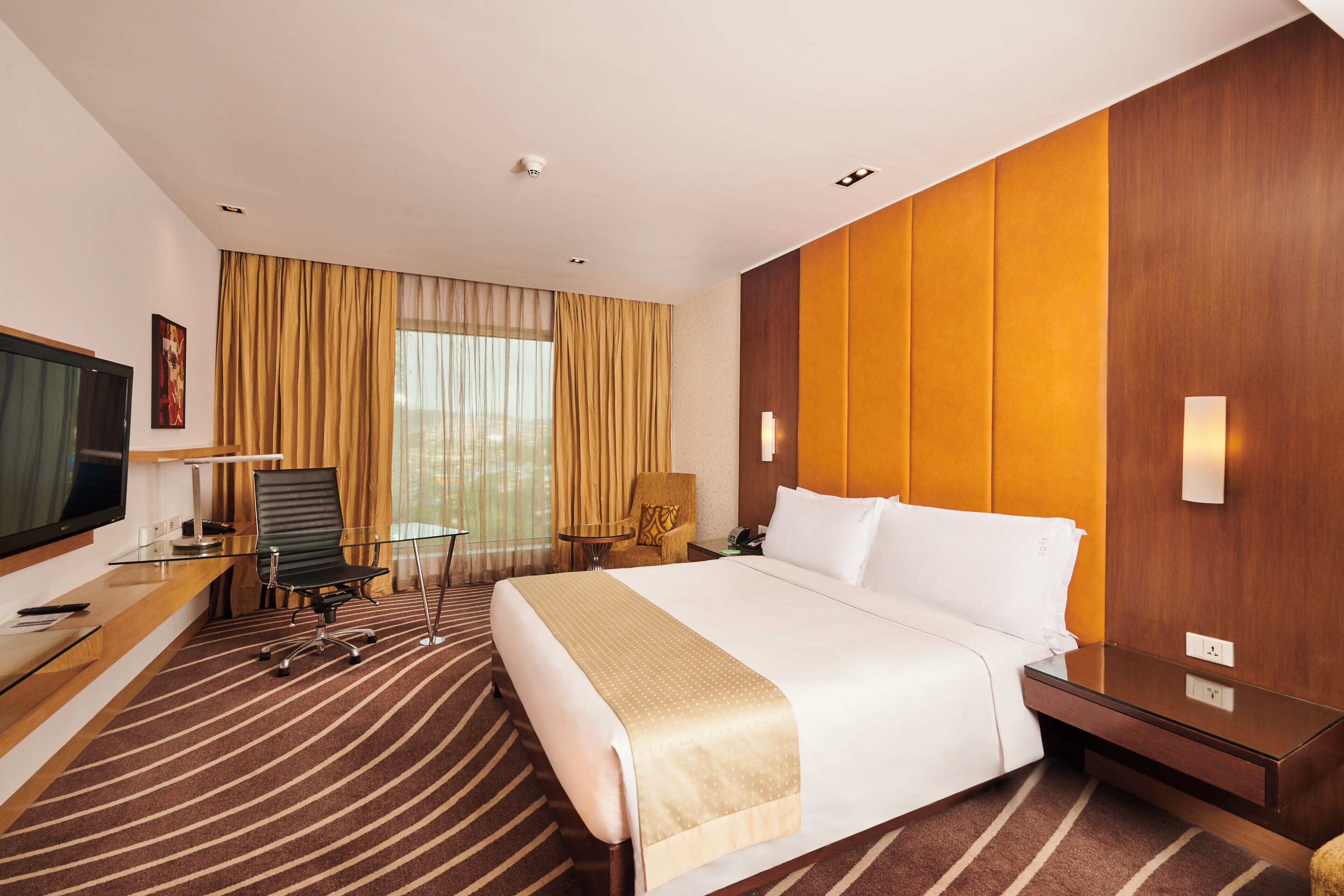 A hotel room with a large bed, a desk with a computer, a chair, a television, and a window with curtains.