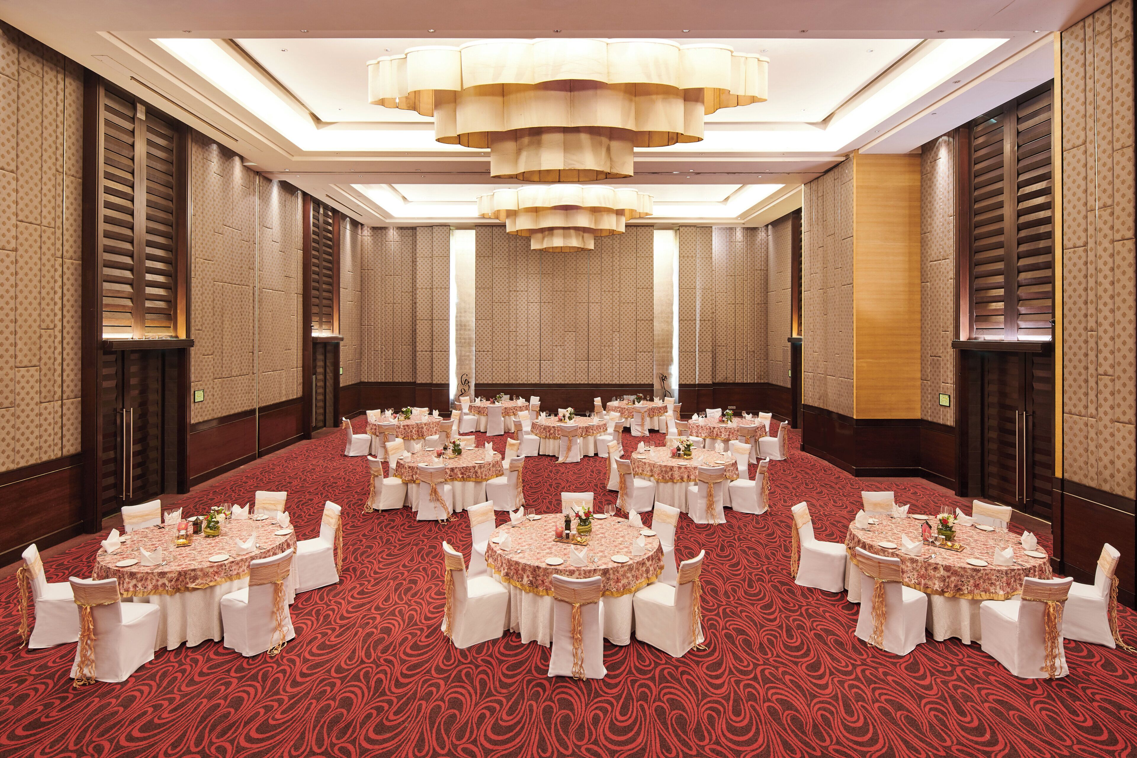 Un amplio salón de banquetes con mesas redondas dispuestas para eventos, que cuenta con una alfombra roja estampada y una lámpara de araña moderna.