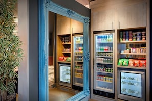 A room with a large mirror, a potted plant, and a minibar stocked with beverages and snacks.