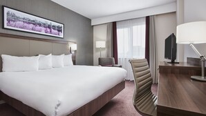 A hotel room with a large bed, a desk with a TV, a chair, and a wall-mounted lamp.