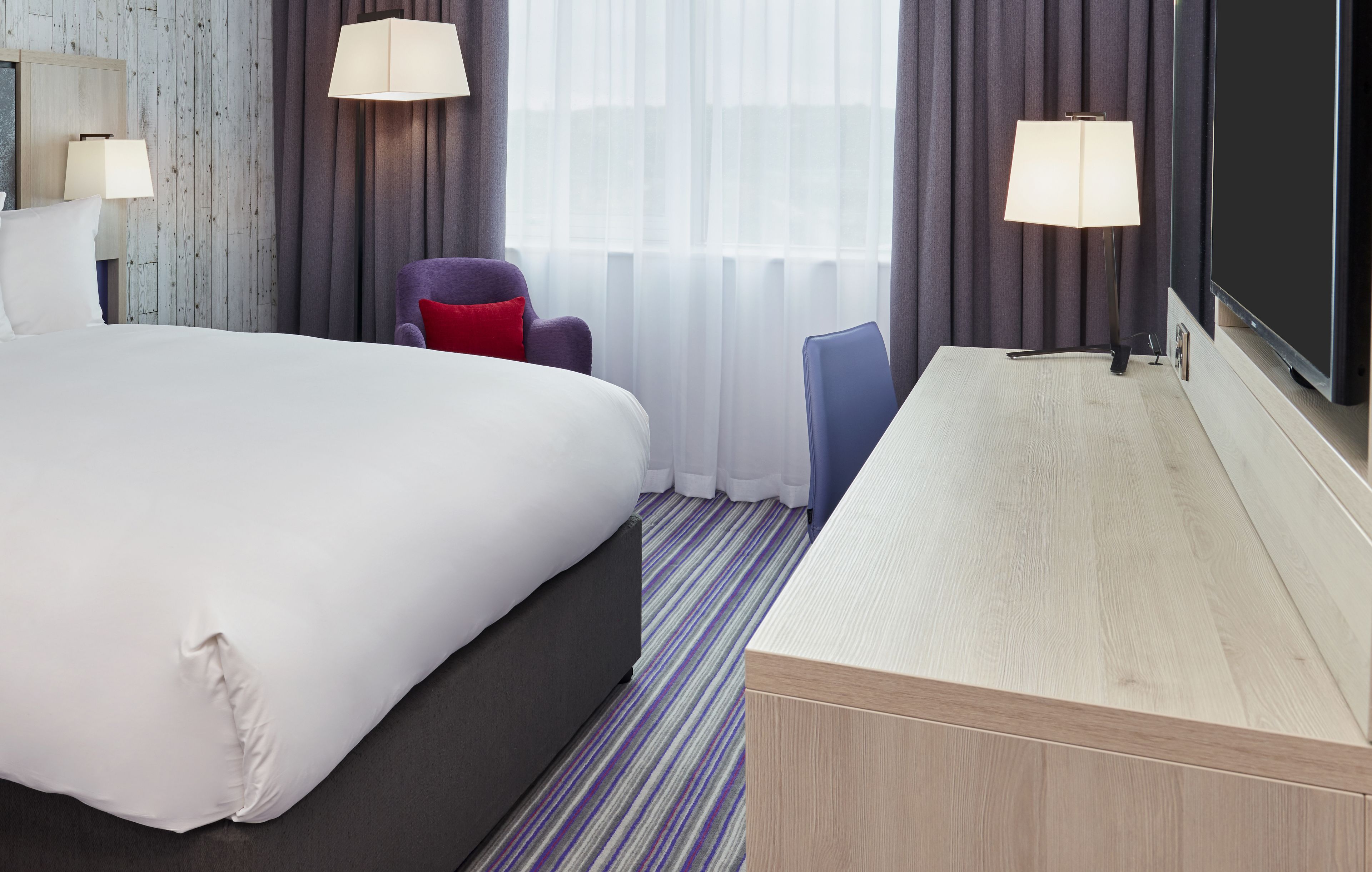 A hotel room with a large bed, a desk, two lamps, a chair, and a television.