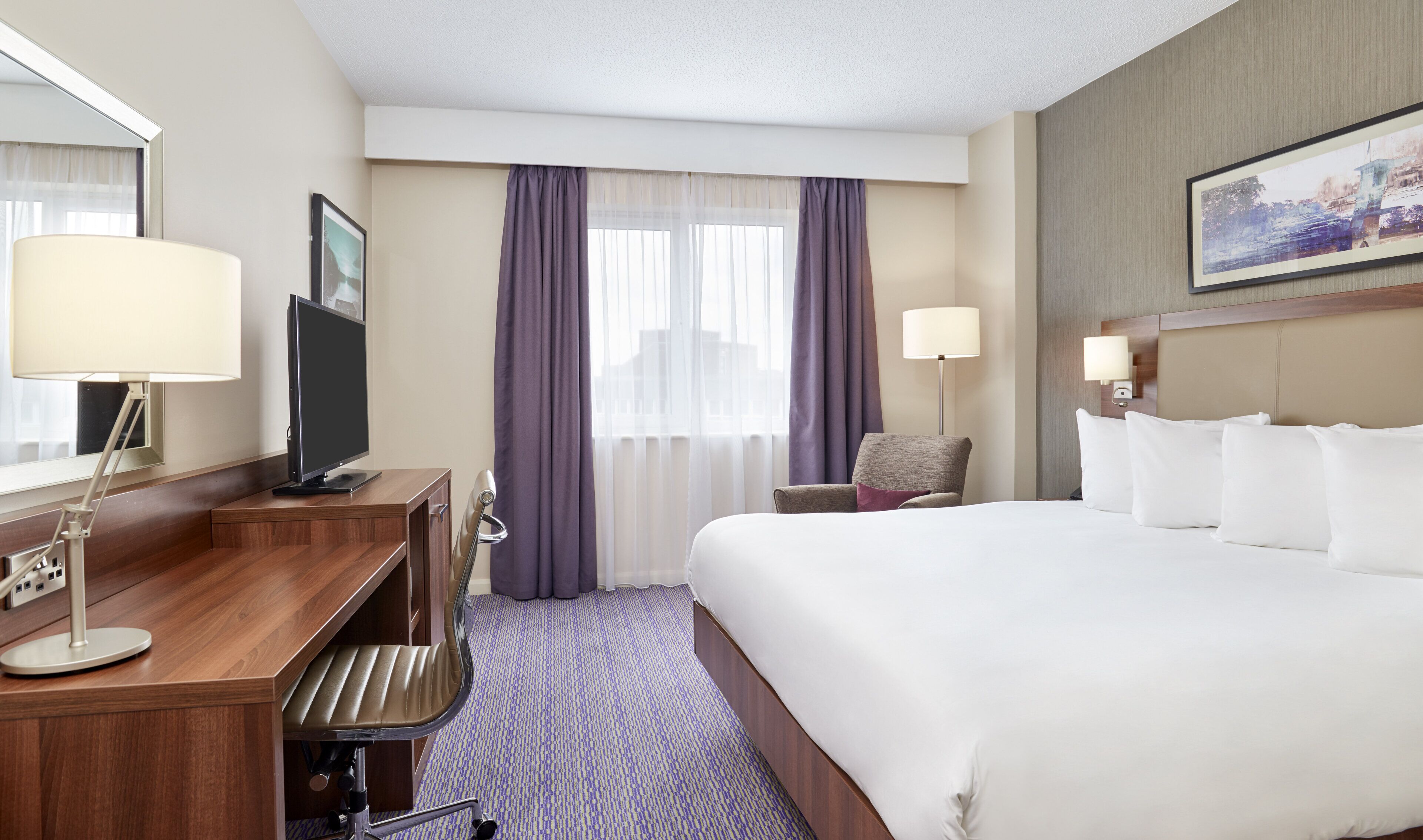 A hotel room with a bed, desk, chair, and a large window with curtains.