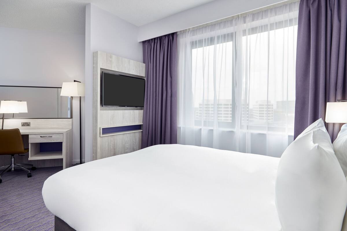 A hotel room with a large bed, a desk with a chair, a flat-screen TV, and a window with purple curtains.
