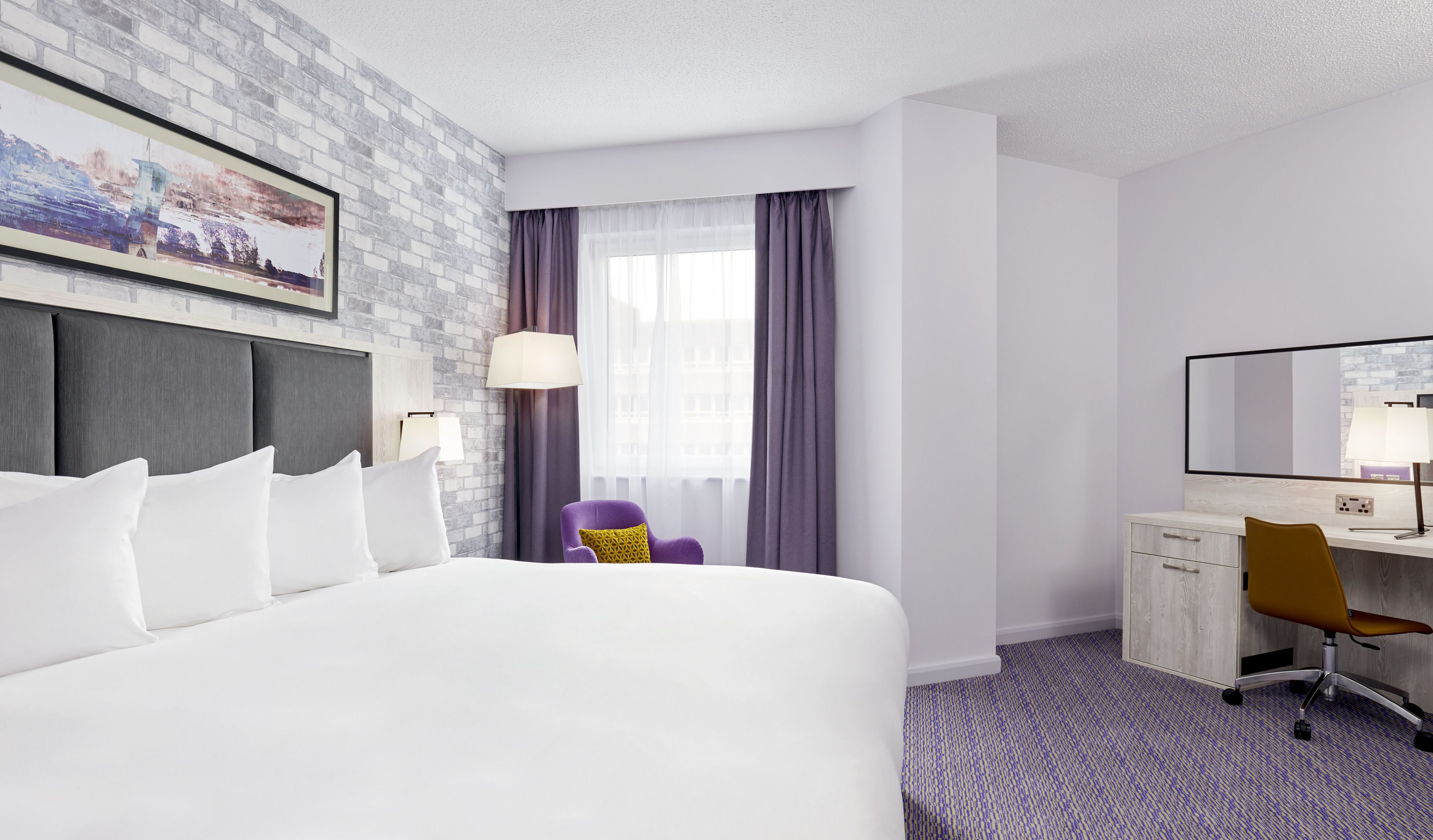 Quarto de hotel com uma cama grande, uma mesa, uma cadeira, uma luminária e uma janela com cortinas.
