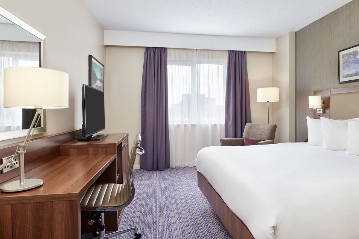 A hotel room with a bed, a desk with a computer, a chair, a lamp, and a window with curtains.