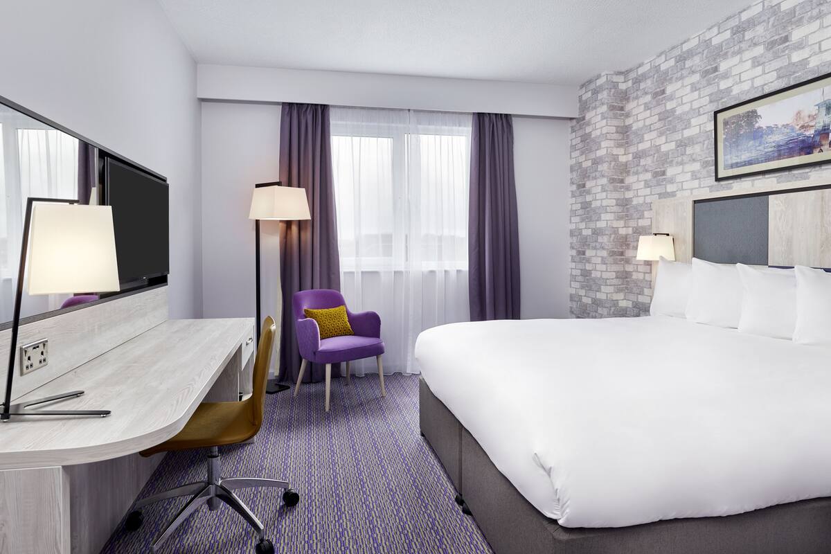 A hotel room with a large bed, a desk with a computer, a chair, and a window with curtains.