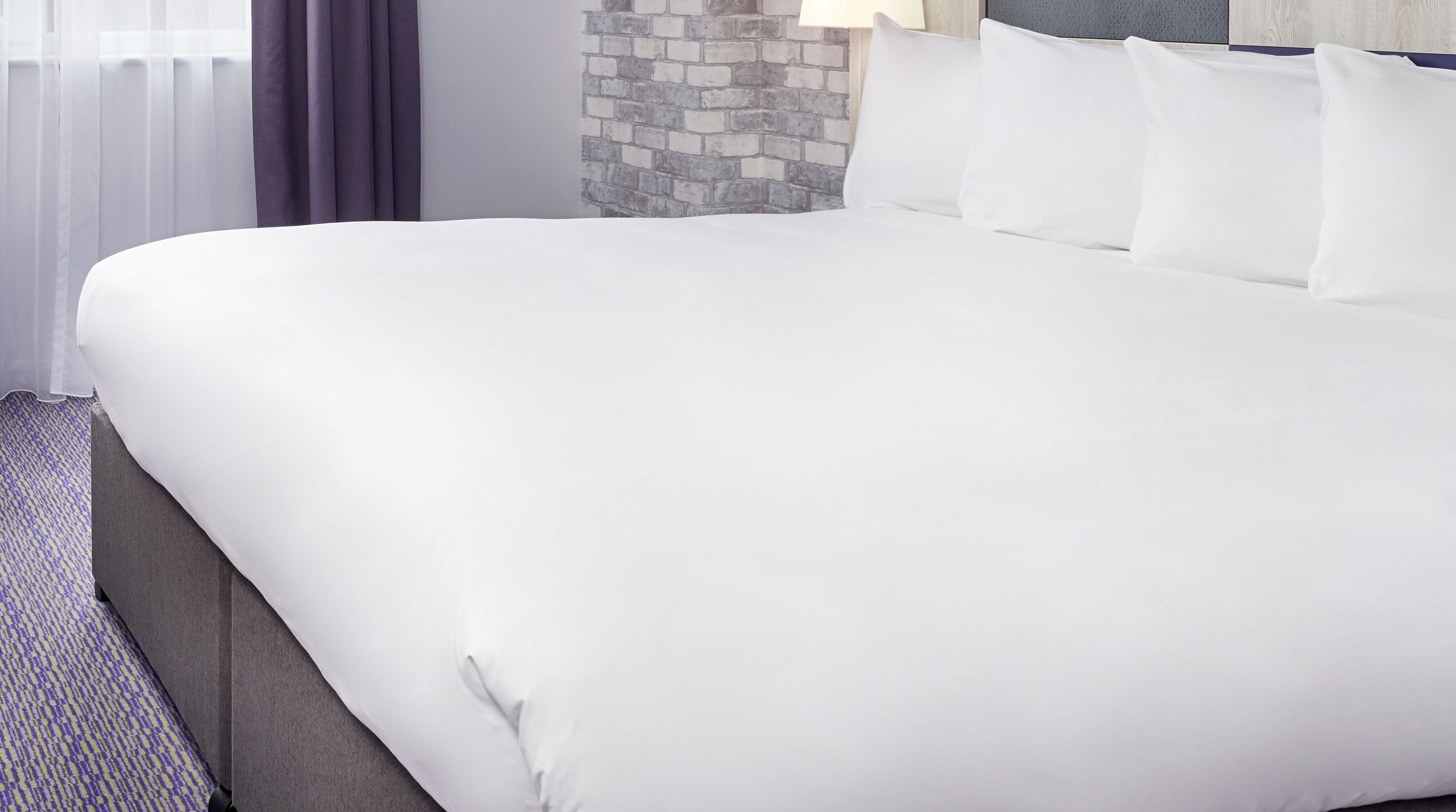 A large bed with white linens, a headboard with a blue accent, and a brick-patterned wall behind it.