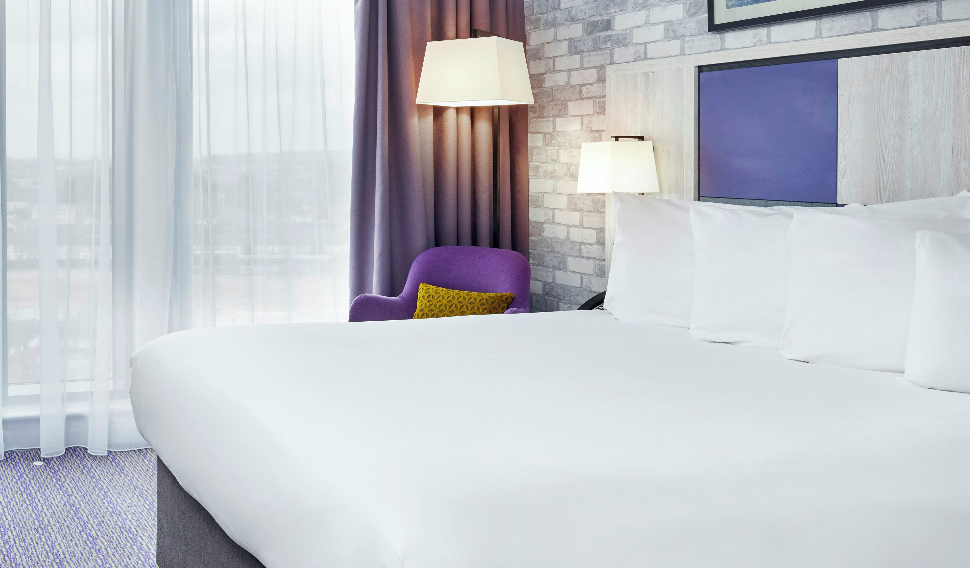 A hotel room with a large bed, a purple armchair, two bedside lamps, and a city view through sheer curtains.