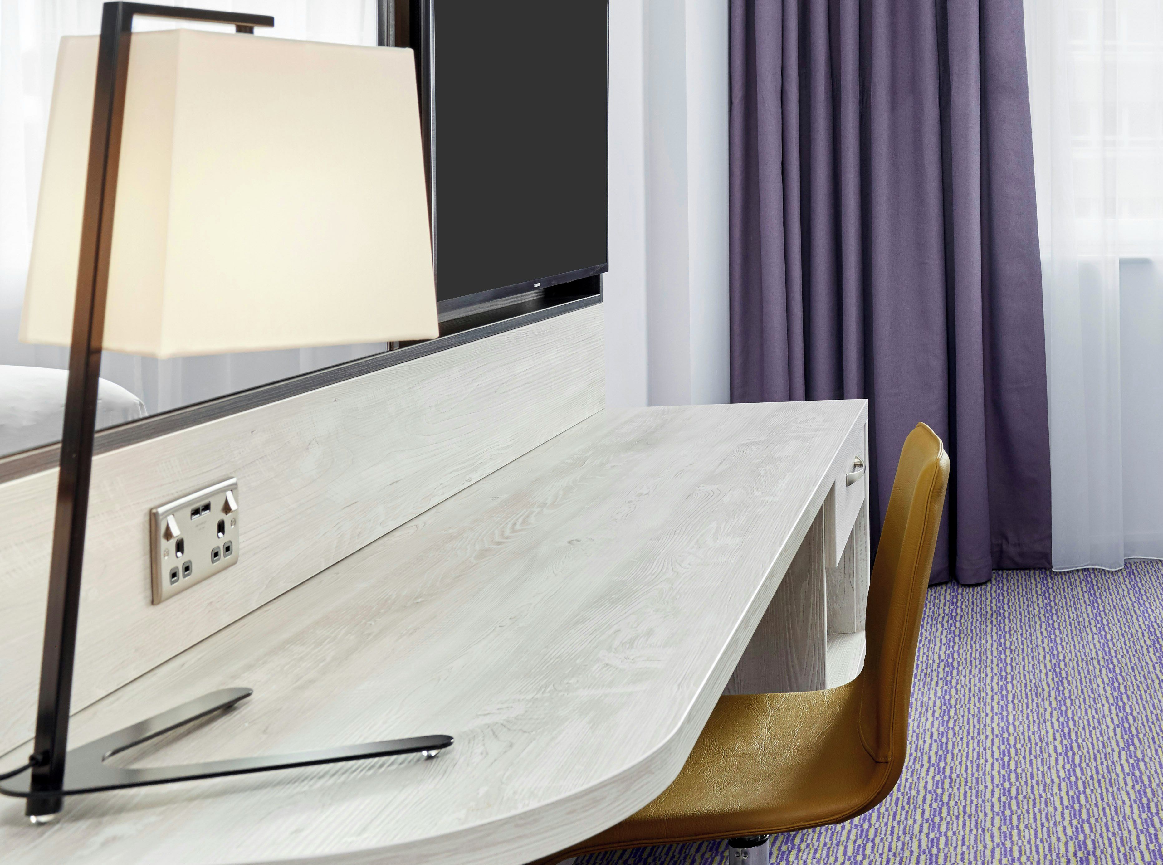 Une chambre d’hôtel équipée d’un bureau, d’une lampe, d’une chaise et d’une fenêtre avec des rideaux.