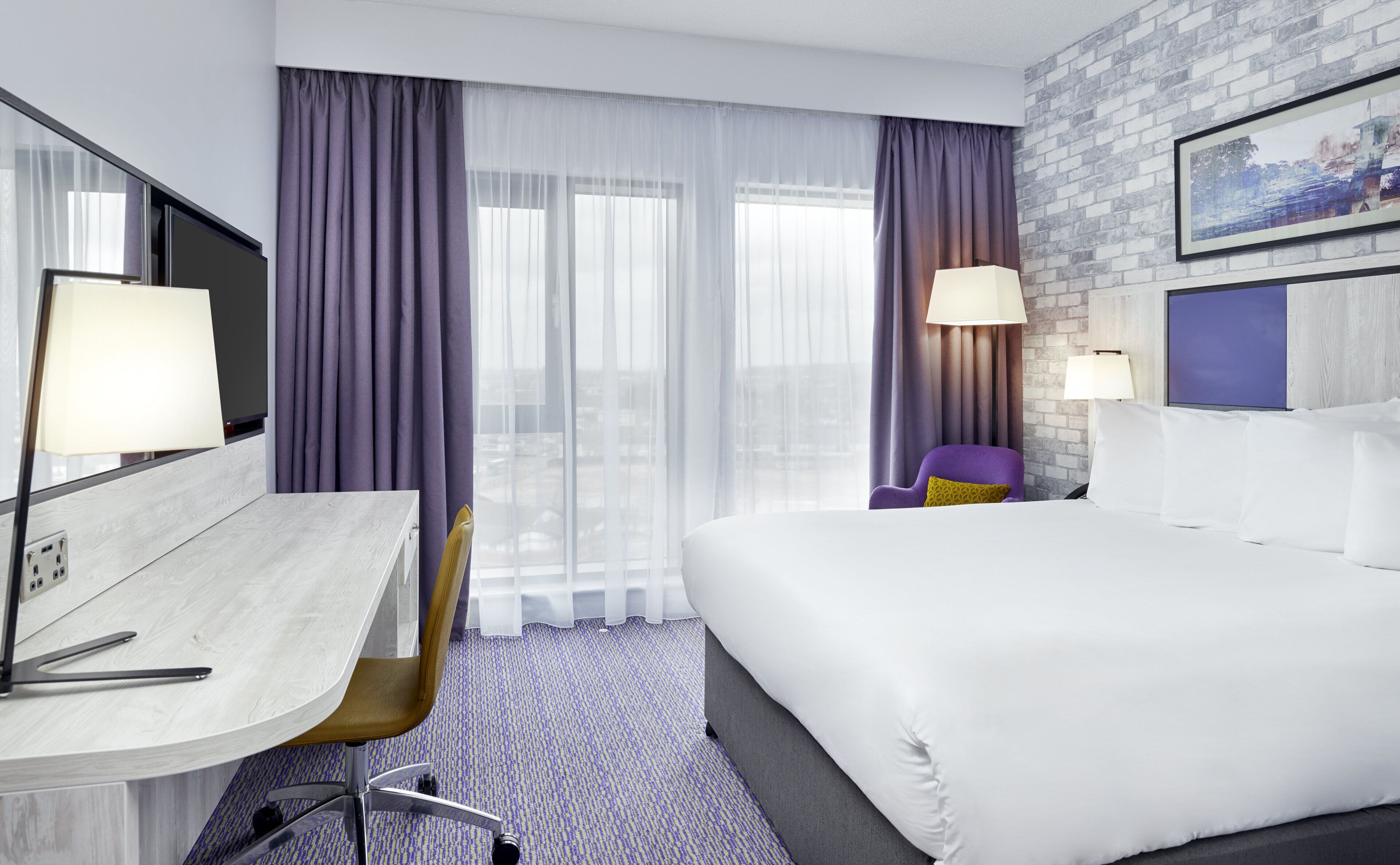 A hotel room with a bed, a desk, a chair, two lamps, and a window with curtains.