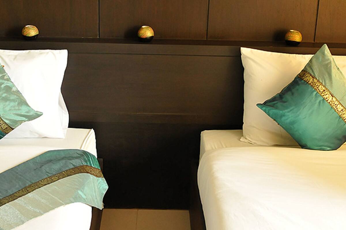 Two beds with white linens and teal pillows with gold trim, set against a dark headboard with three gold knobs.