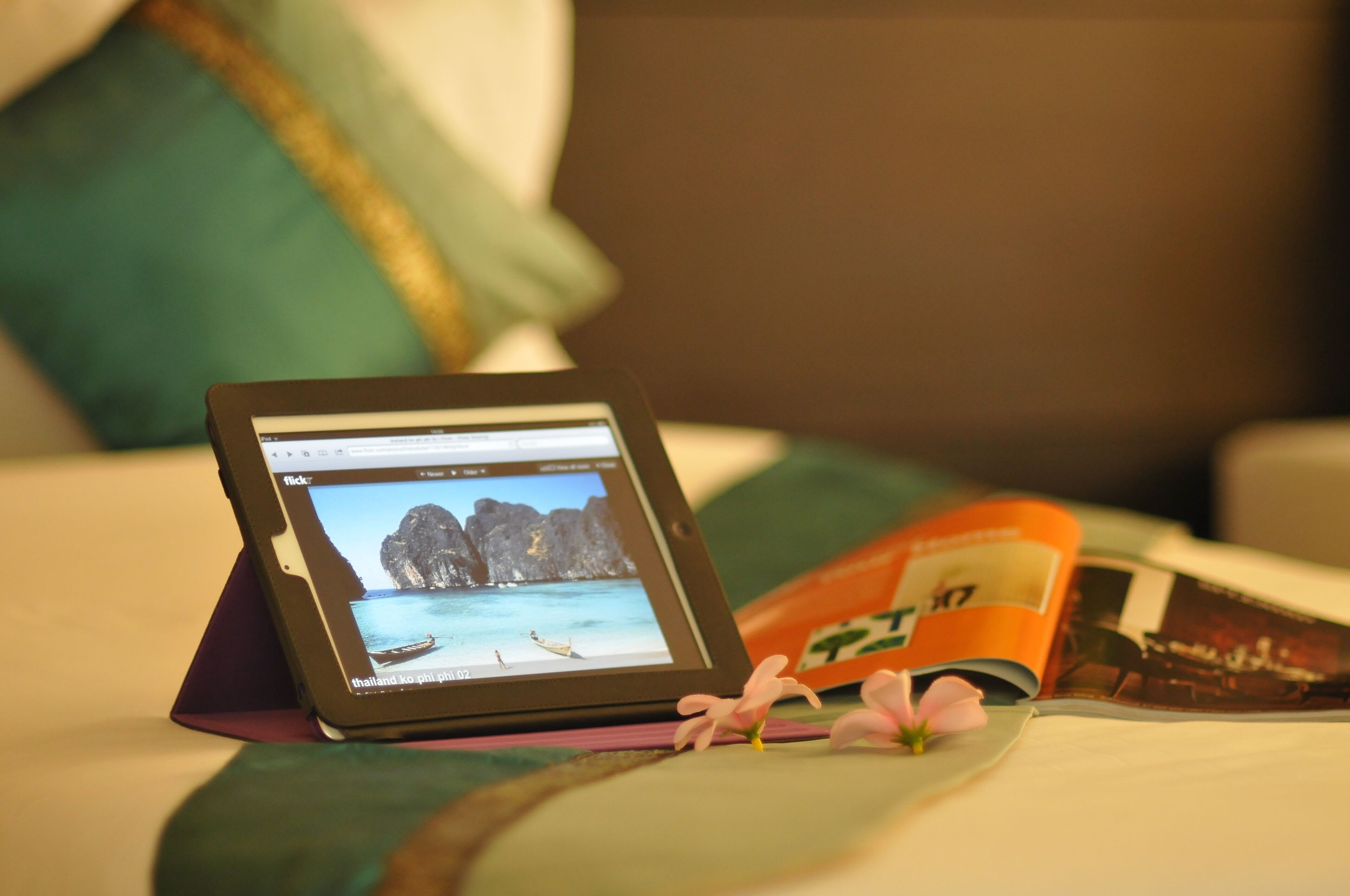 A tablet displaying a travel website on a bed with a magazine and a pillow.