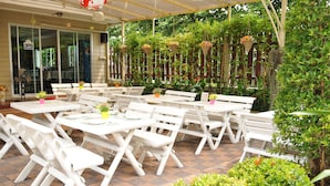 Une terrasse extérieure avec des tables et des chaises blanches, entourée de verdure et agrémentée de jardinières suspendues.