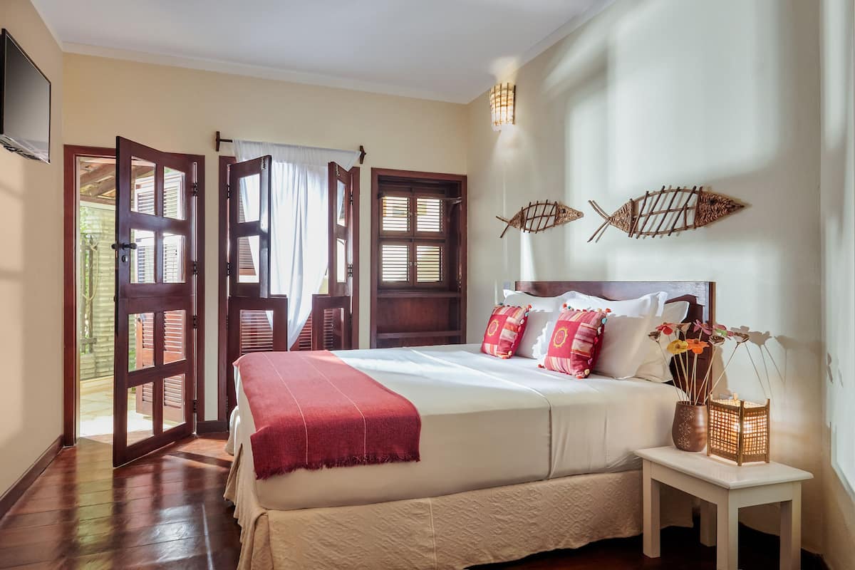Quarto com cama grande, mesa de cabeceira pequena, televisão na parede e janela com persianas.