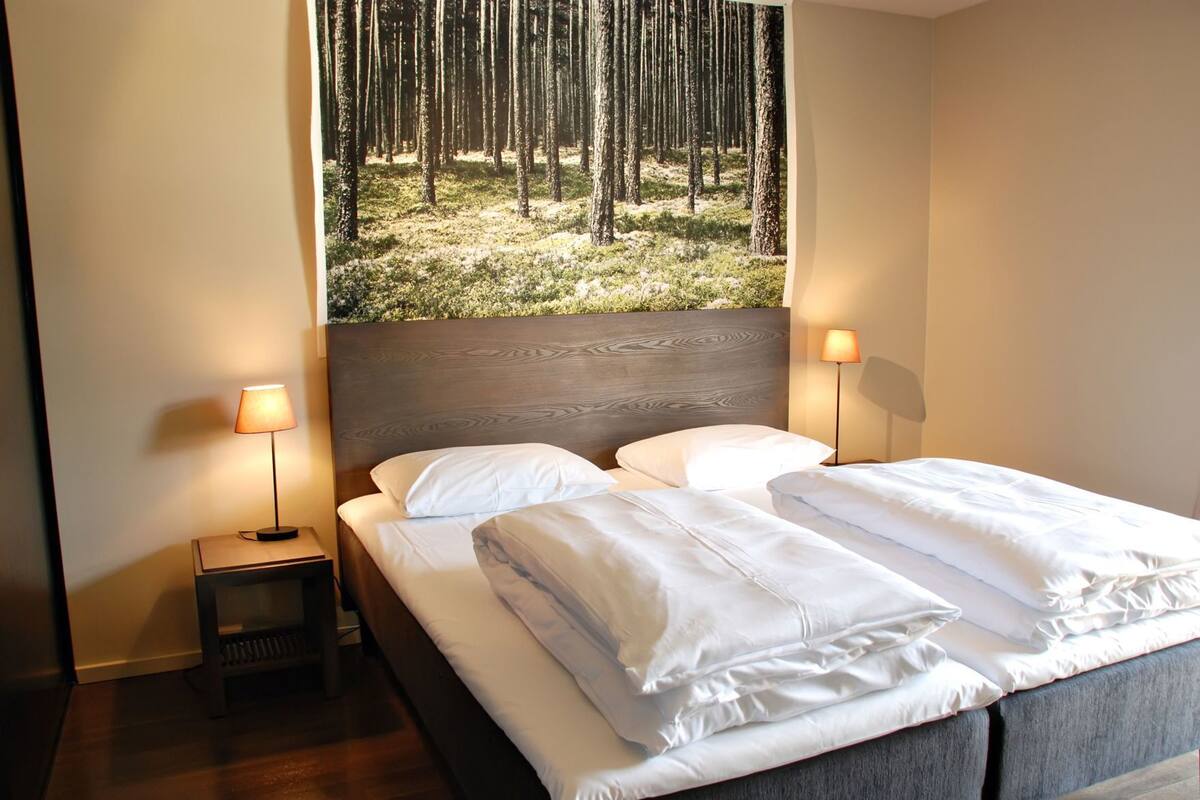 A hotel room with a bed, two bedside lamps, a nightstand, and a large forest view.