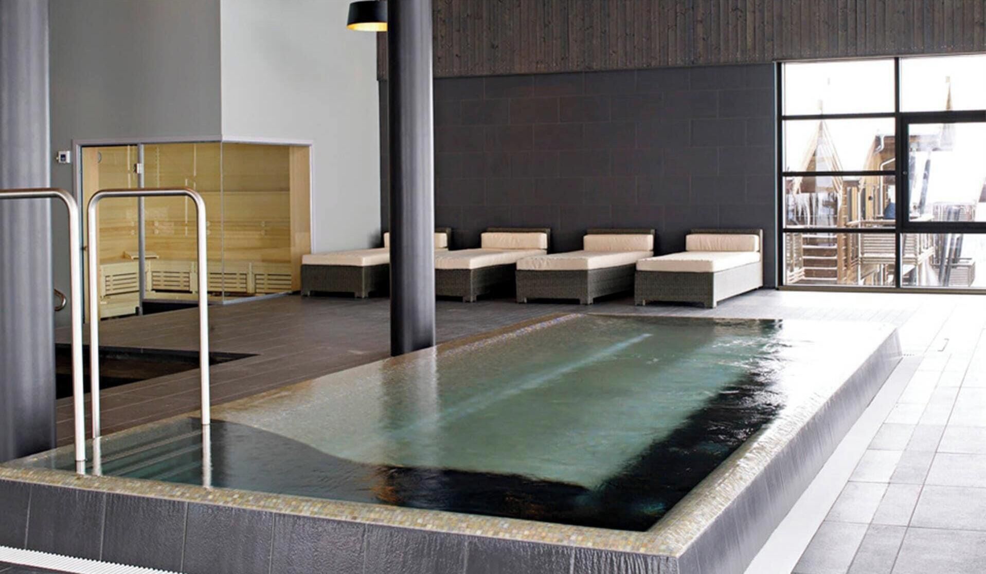A modern indoor swimming pool with a glass barrier, seating area, and a view of the cityscape through large windows.