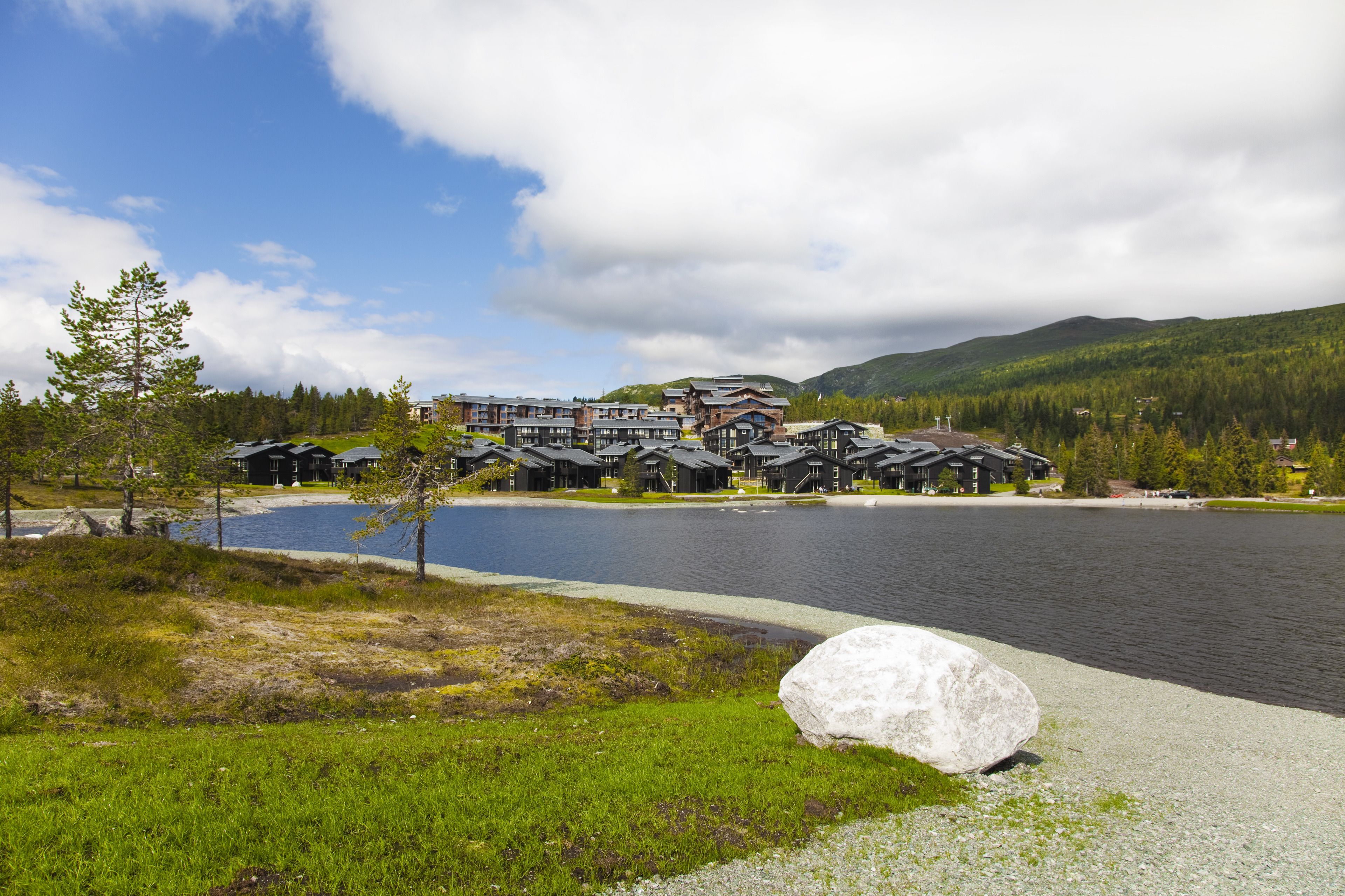 Un hotel frente al lago con varias construcciones de estilo chalet, una gran roca en primer plano y montañas al fondo.