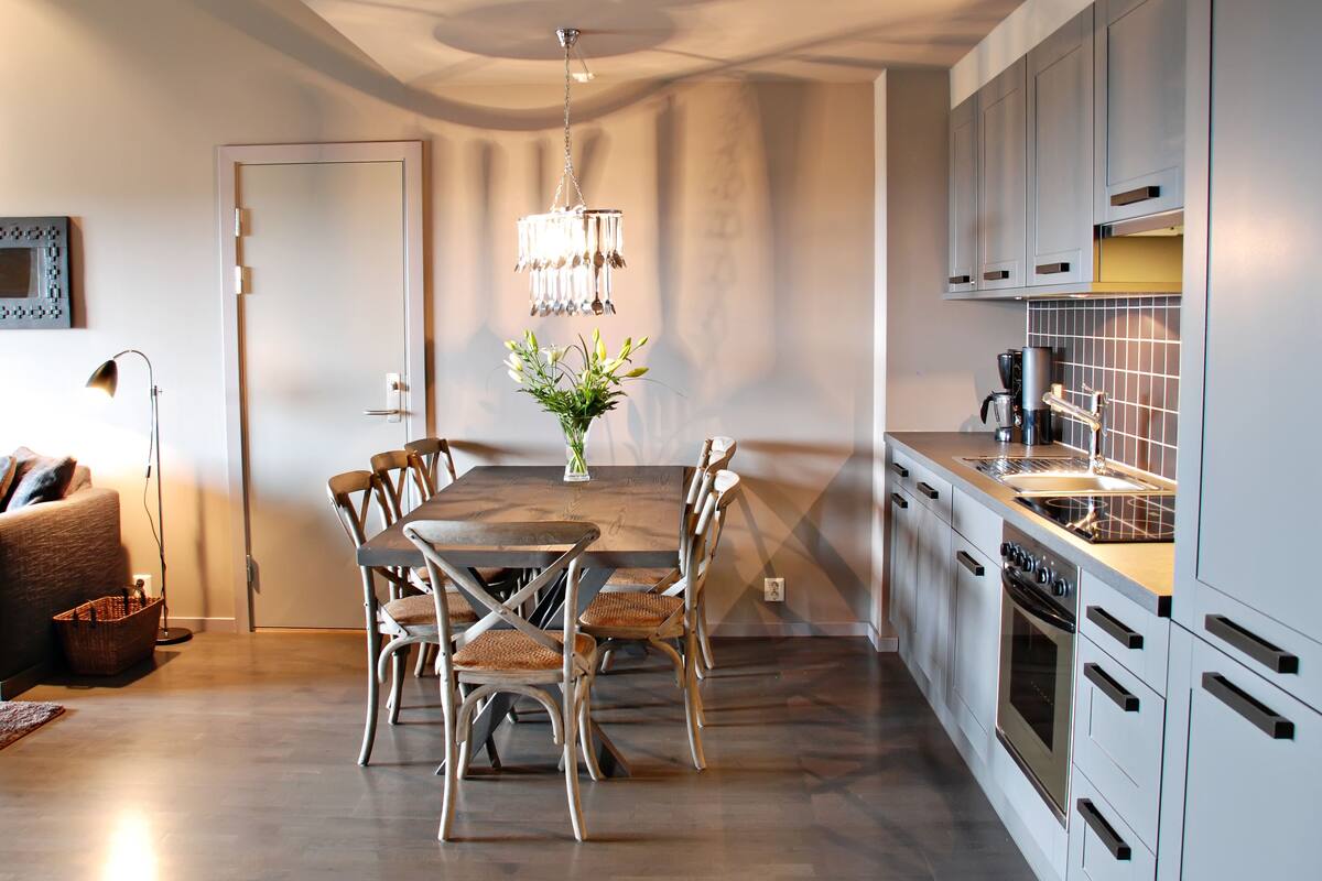 A modern kitchen with a dining area, featuring a wooden table, chairs, a chandelier, and built-in appliances.