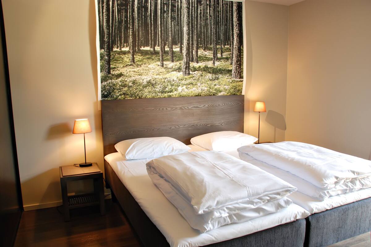 A neatly made bed with white linens and pillows, a small bedside table with a lamp, and a large window with a forest view.
