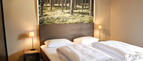 A neatly made bed with white linens and pillows, a small bedside table with a lamp, and a large window with a forest view.